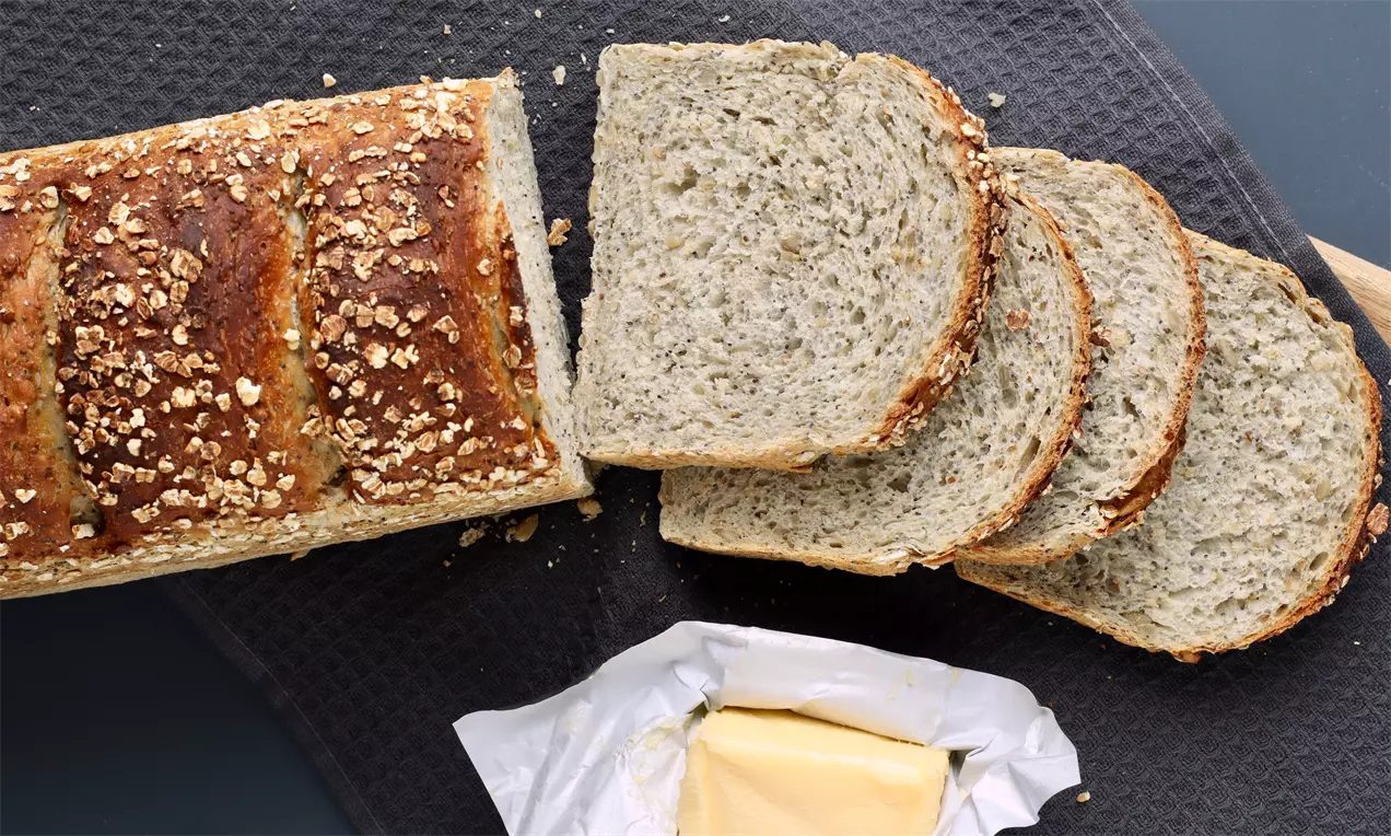 Firkantet gyllenbrunt kernebrød med sprø skorpe og havre på toppen, skåret i skiver ved siden av smør på papir, liggende på mørkt underlag