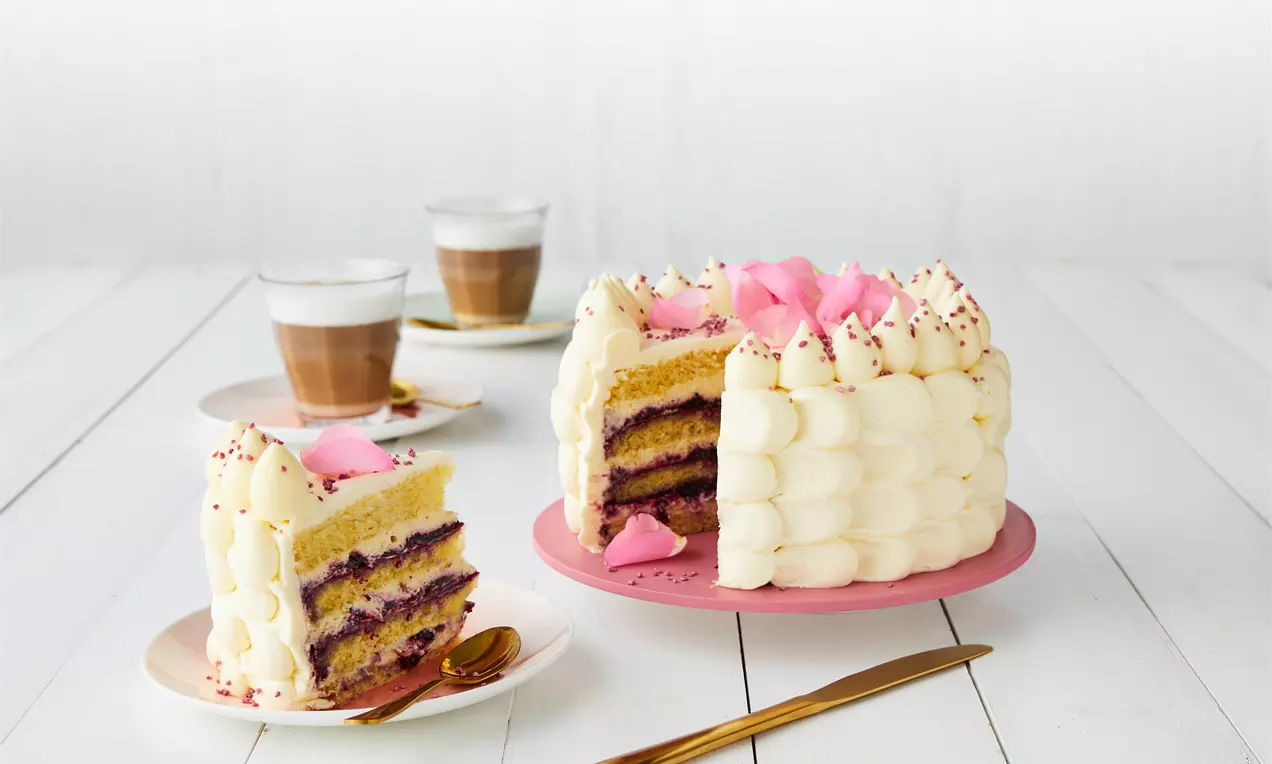 Høy lagkake med lys vaniljekrem og syltetøy i lilla lag, dekorert med rosa kremtopper og blomster på rosa fat og hvitt bord