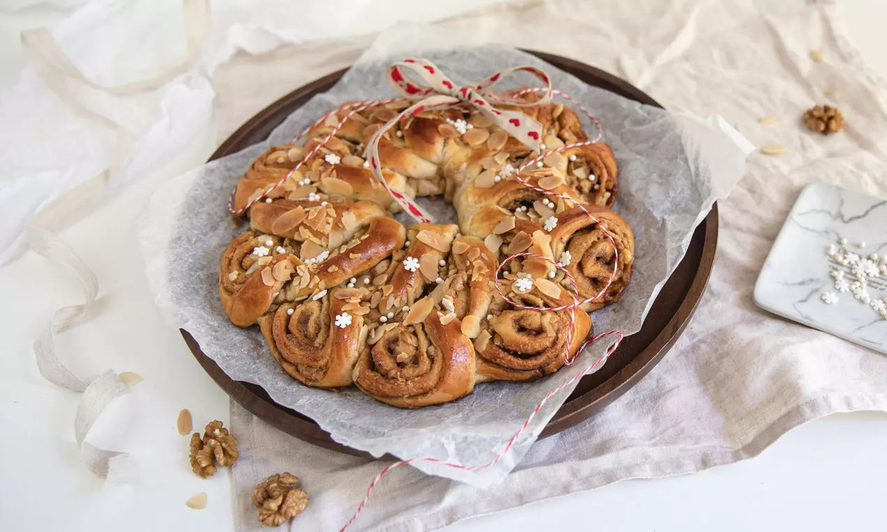 Kanel-valnøddekringle i gyllenbrune spiraler toppet med snøfnuggpynt og mandelskiver på bakepapir, knyttet med rødhvit bånd