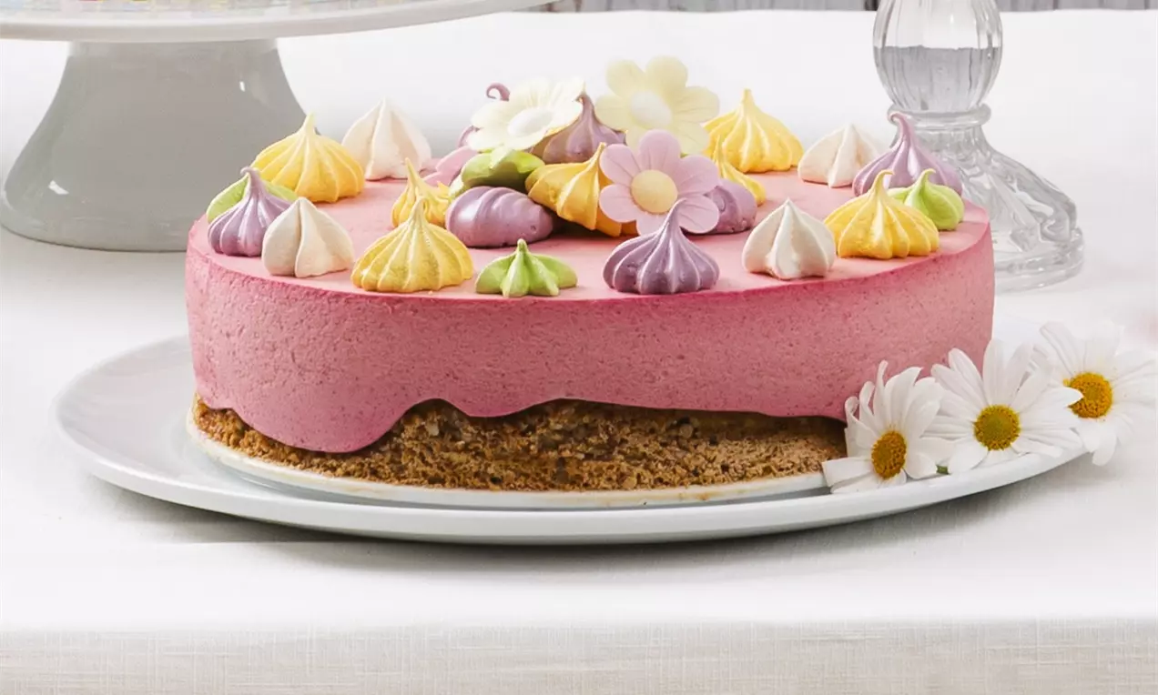 Rosa bringebærostekake på en brun nøttebunn, toppet med små, fargerike marengstopper og blomster på et hvitt fat med prestekrager ved siden av