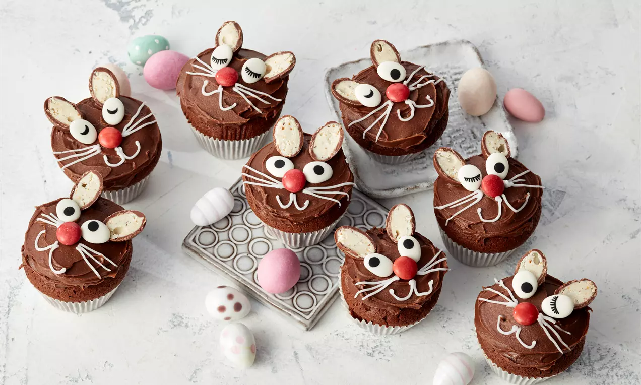 Sjokolademuffins dekorert som kaniner med hvite og røde detaljer arrangert på et lyst bord med små fargede påskeegg rundt