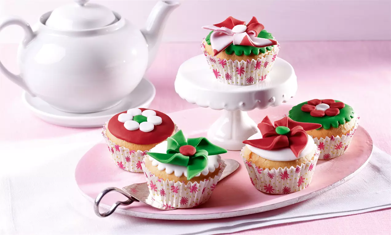 Muffins i lyse bakeformer med rødt, grønt og hvitt sukkertøyblomstertrekk på et rosa fat og et hvitt kakefat ved siden av en hvit tekanne
