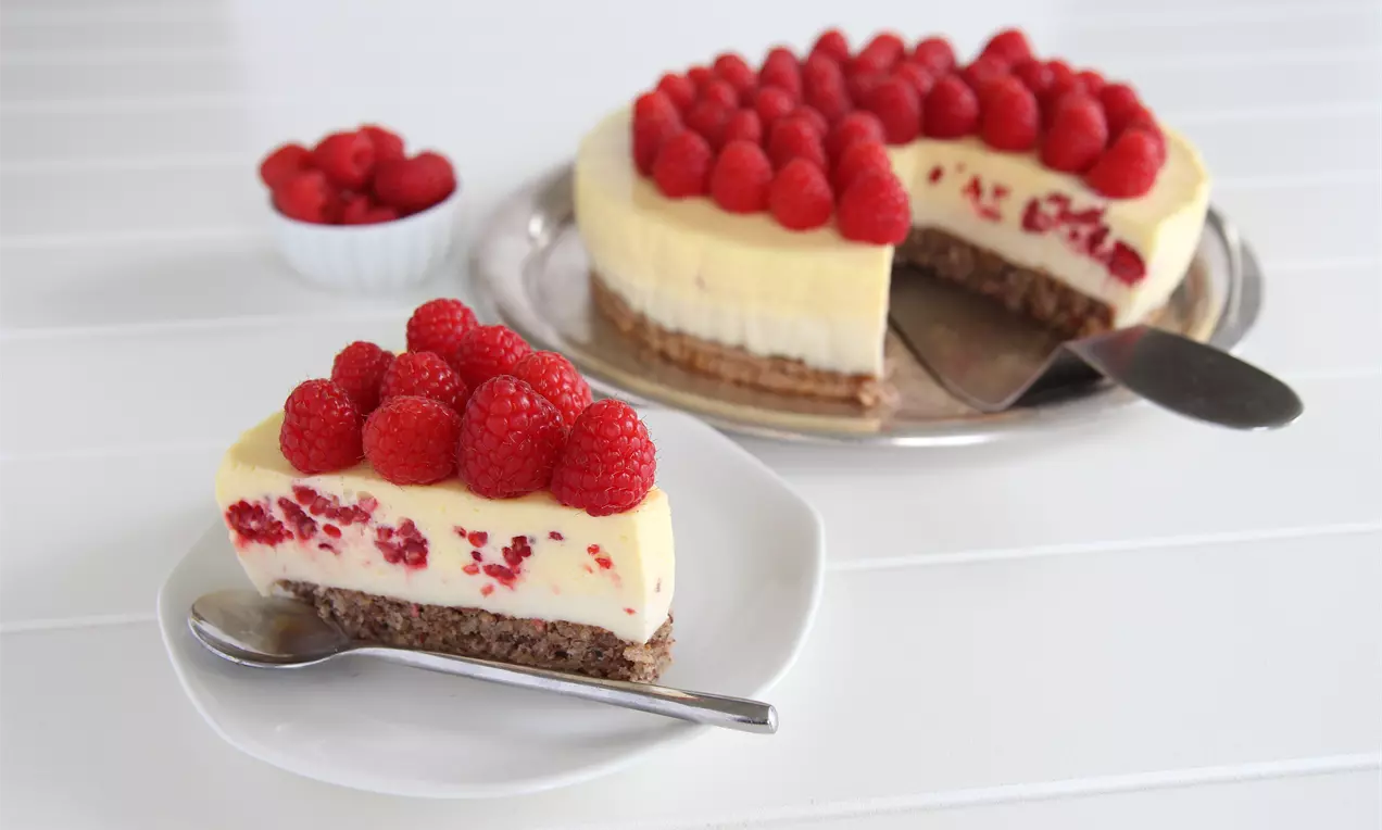 Moussekake med lys kremfarget pannacotta og bringebær på brun bunn, en servering på hvit tallerken med sølvskje, og en kake på serveringsfat