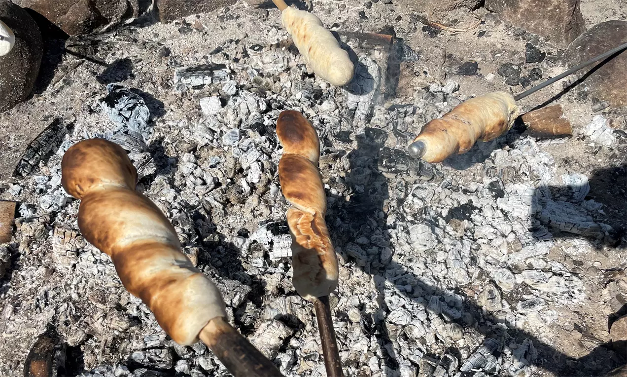Snobrød brunsprø og gyldne på trekkerter, vridd rundt pinnene over glødende grå aske og kull med steiner i bakgrunnen