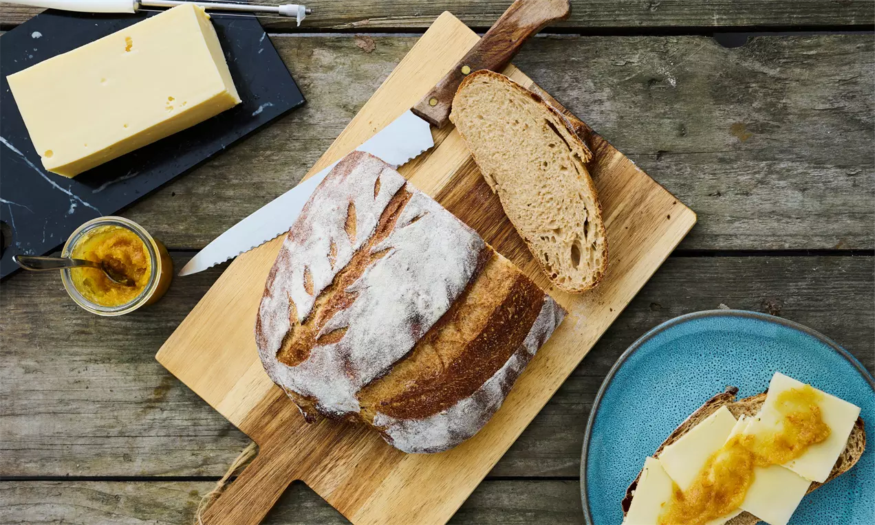 Surdeigsbrød med gyllen og sprukket skorpe ligger i en trekantet hevekurv på et bord med skiver, smør, kniv og brødskive med pålegg