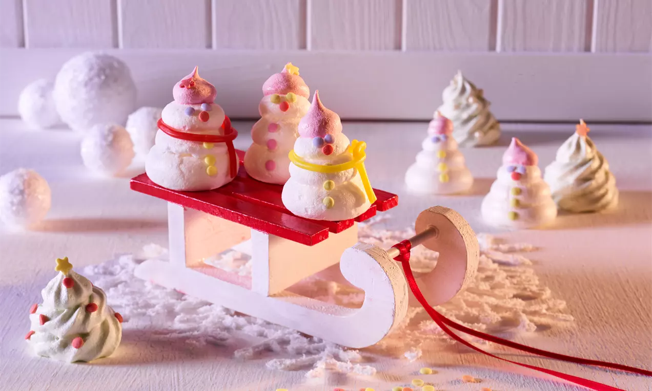 Små hvite marengs formet som snømenn med rosa hatter og som juletrær med stjerner og strø pyntet på en rød kjelke og hvit bakgrunn