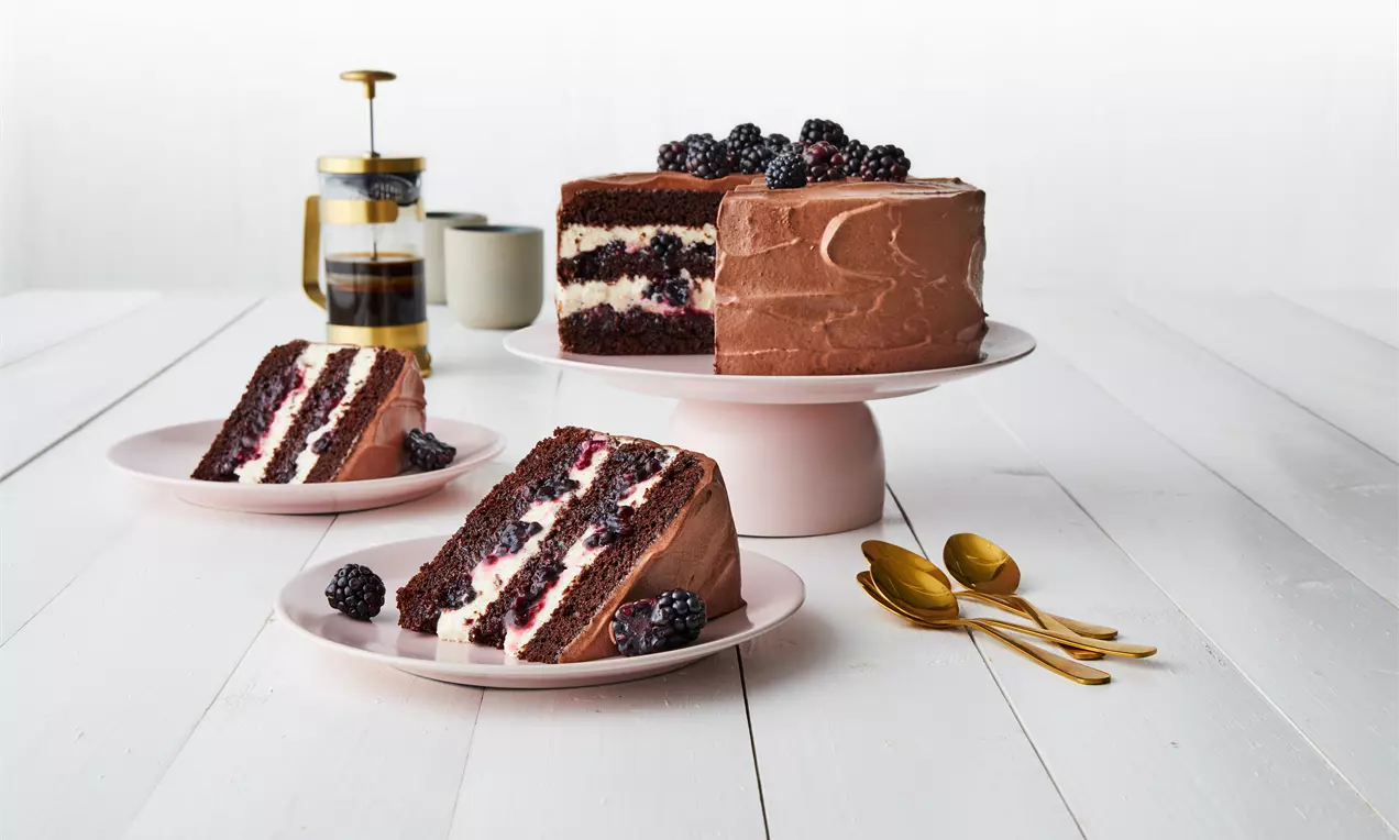 Sjokoladekake med lag av krem og bjørnebær på hvitt kakefat, pyntet med bjørnebær på toppen og serveringsstykker på rosa tallerkener