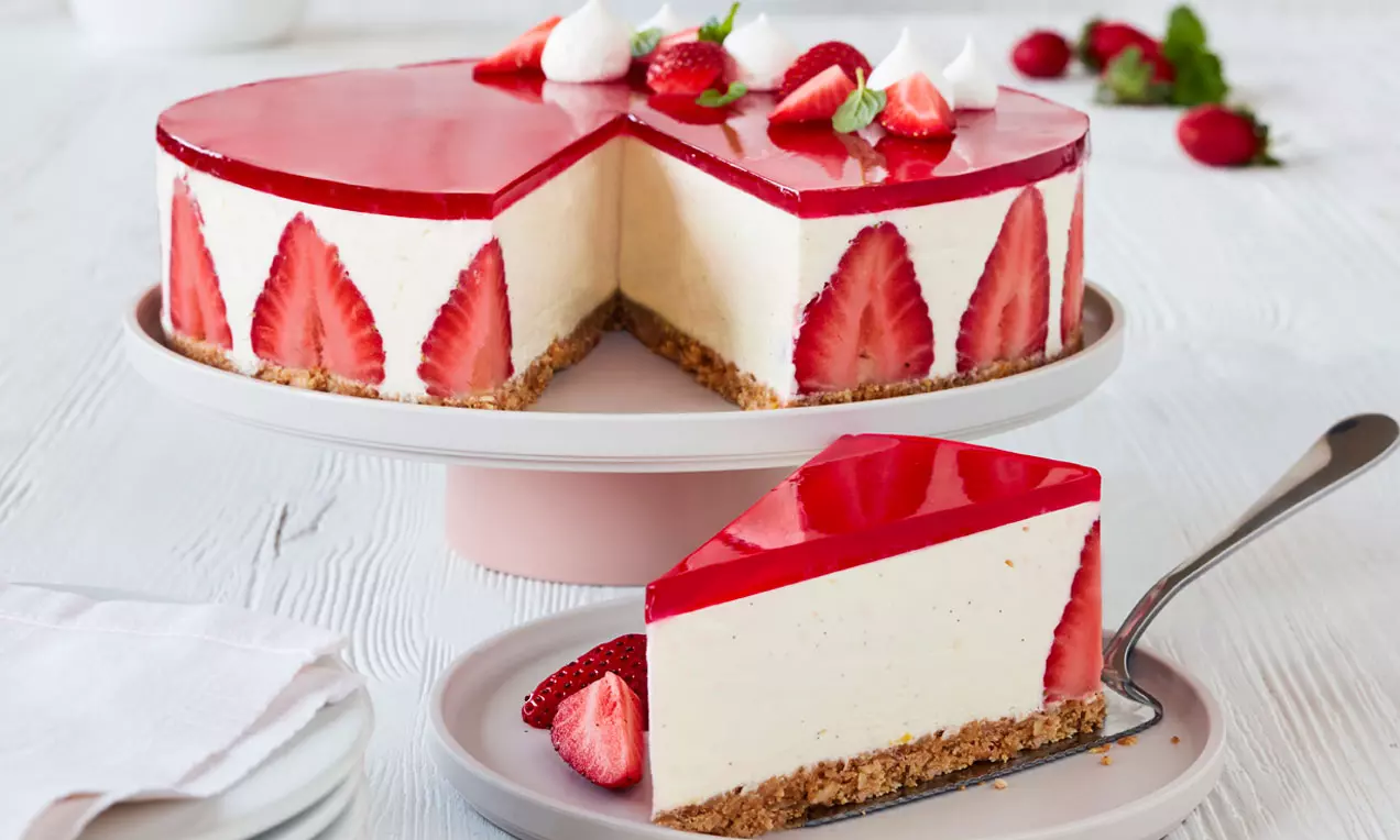 Cheesecake med lys kjeksbunn, kremet vaniljefyll, røde jordbær i kanten, blank jordbærgelé og friske bær på hvitt kakefat