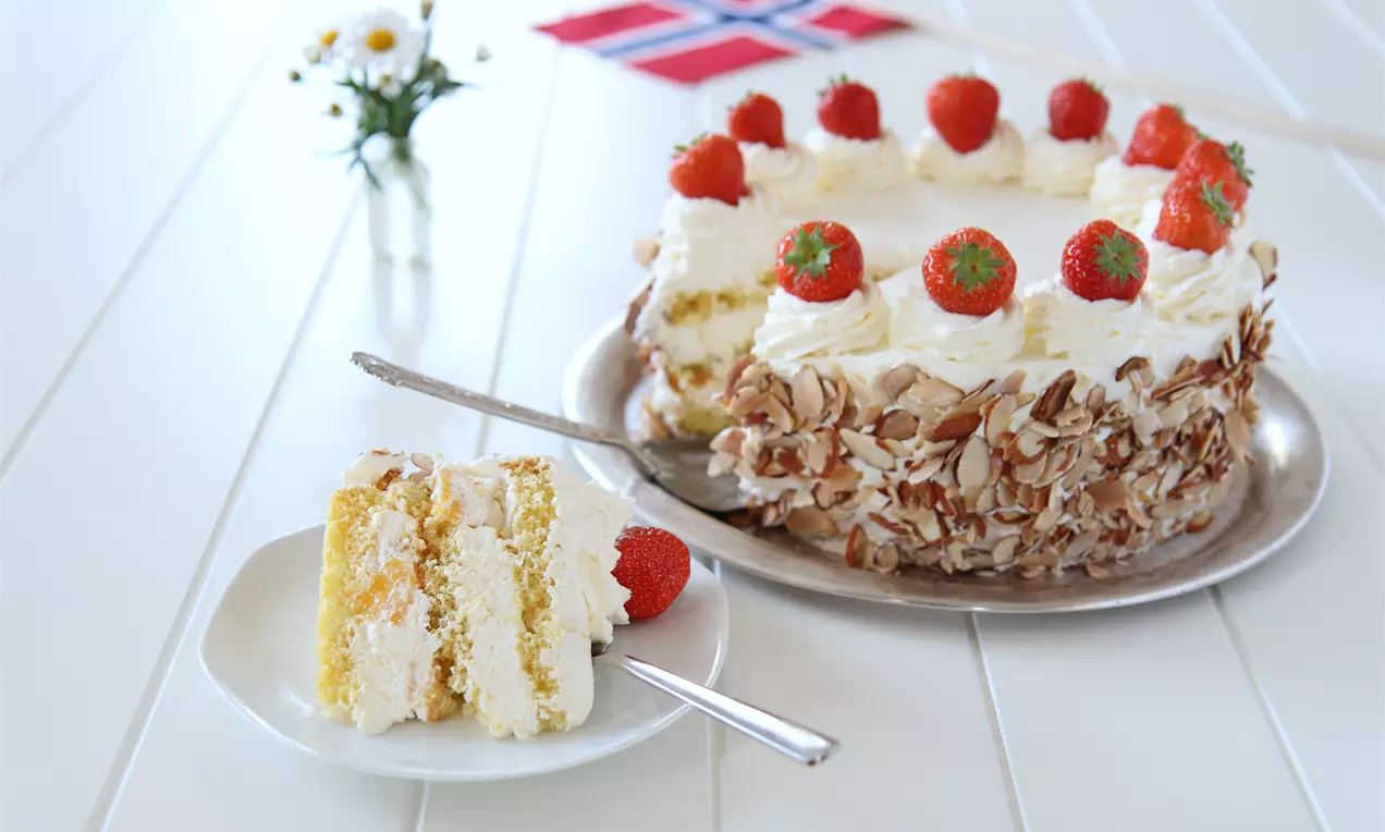 Rund kake med hvit krem, jordbær på toppen og skivede mandler på sidene, servert på et brett ved siden av en kakebit på tallerken