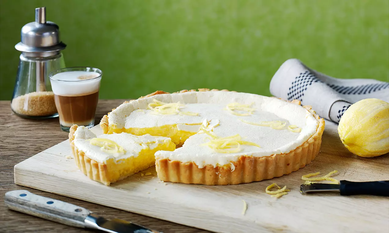 Sitrontarte med gyllen, riflet paiskorpe og lys sitronkrem toppet med revet sitronskall på trefjøl med sitron, kniv og kaffeglass ved siden av